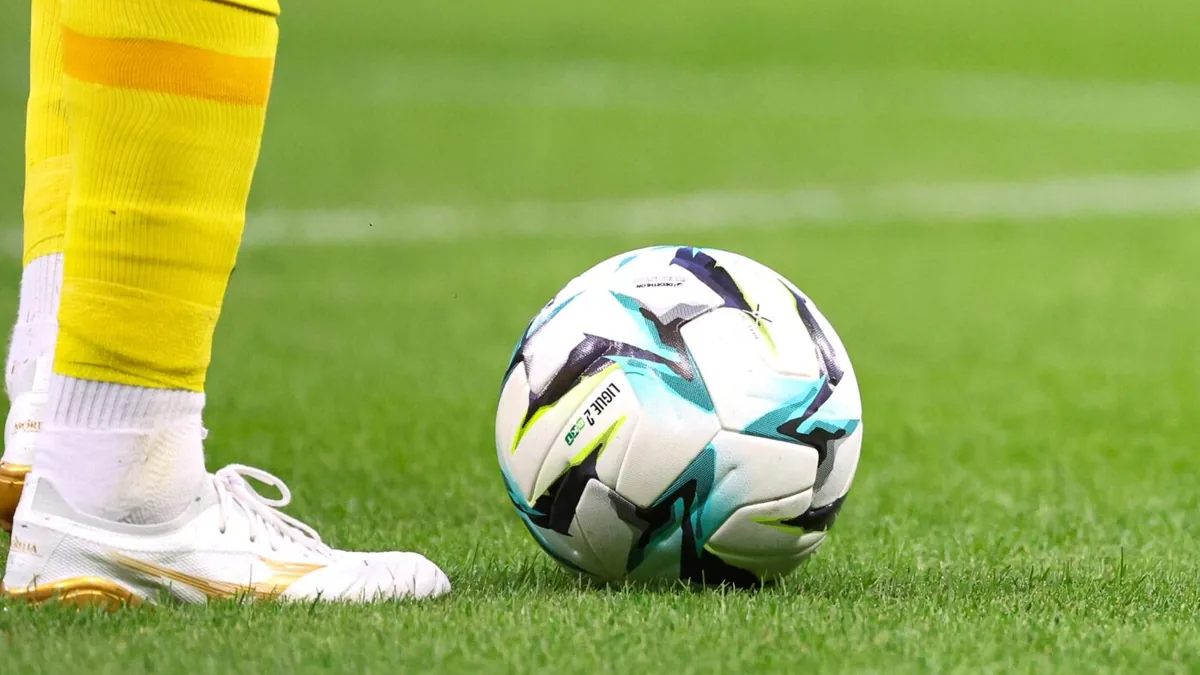 Ballon officiel de la Ligue 2 posé sur la pelouse au pied d'un joueur en chaussettes jaunes
